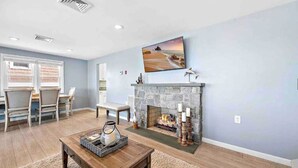 A living room with a stone fireplace, a television mounted above it, and a dining area with chairs and a table. - Sunset Bay LBI lower unit (Long Beach)