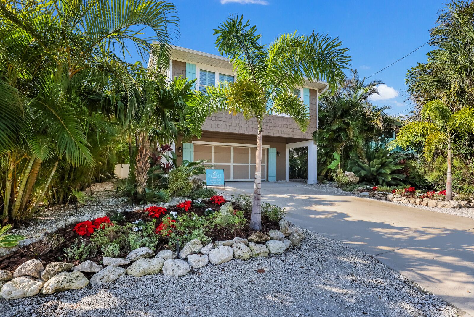 A modern two-story house with a garage, surrounded by tropical plants and a gravel driveway.