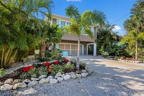A modern two-story house with a garage, surrounded by tropical plants and a gravel driveway.
