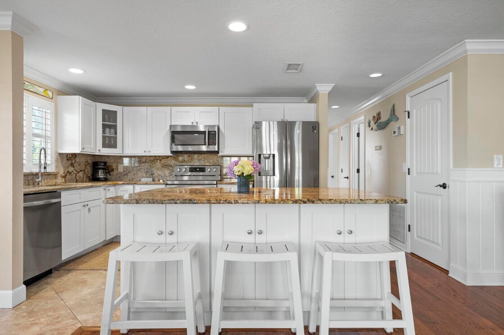 A kitchen with white cabinets, a granite countertop, stainless steel appliances, and a bar area with white stools.