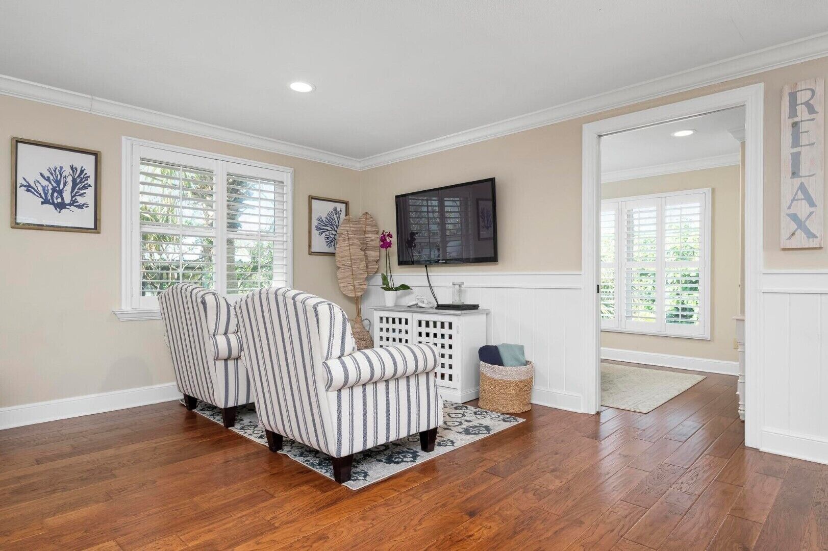 Une pièce avec un plancher en bois, deux fauteuils à rayures, une console blanche et un téléviseur fixé au mur.