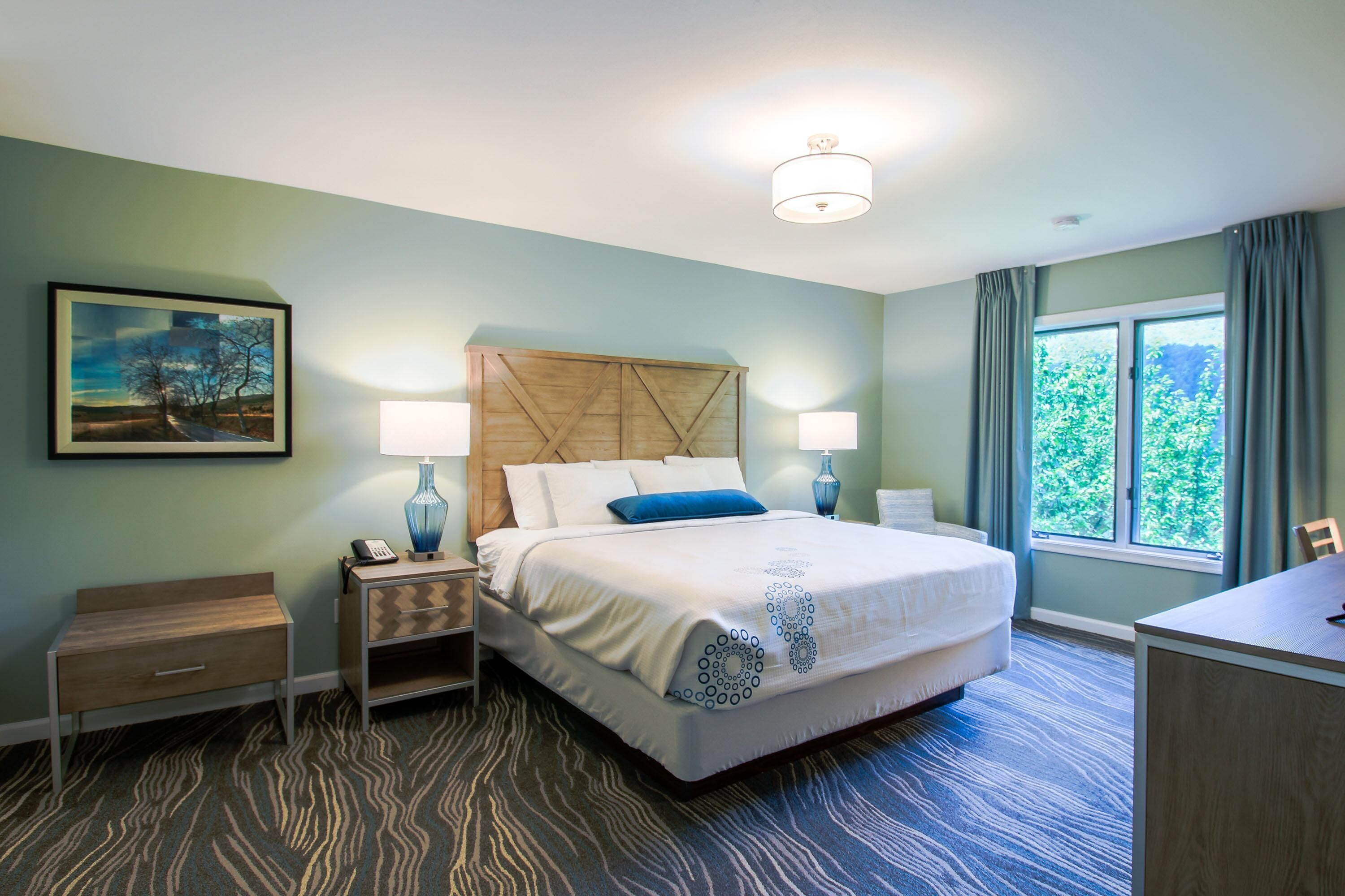 A hotel room with a large bed, two bedside tables, a desk, a chair, a window with curtains, and a painting on the wall.