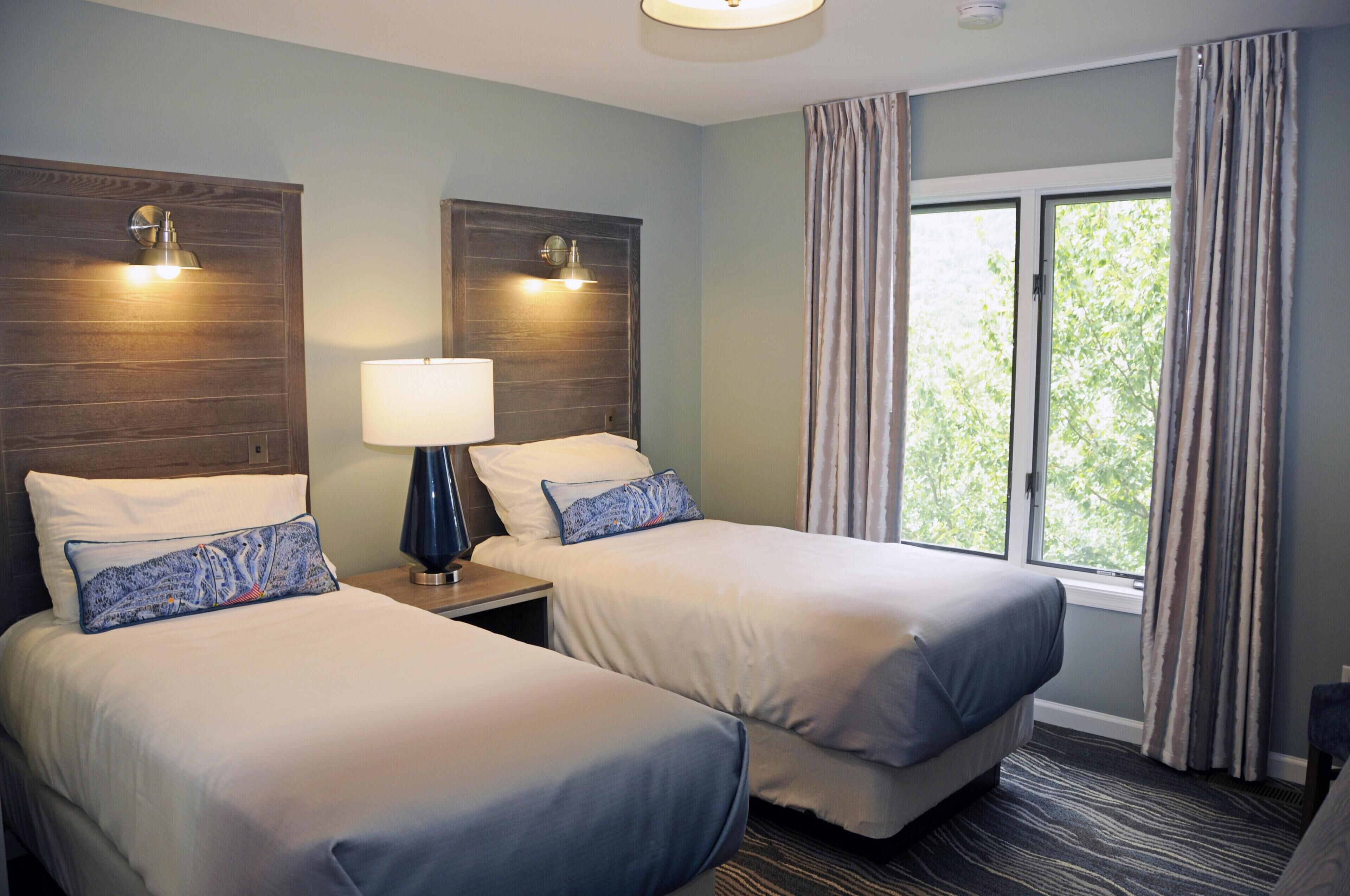 A hotel room with two beds, a nightstand with a lamp, and a window with curtains.