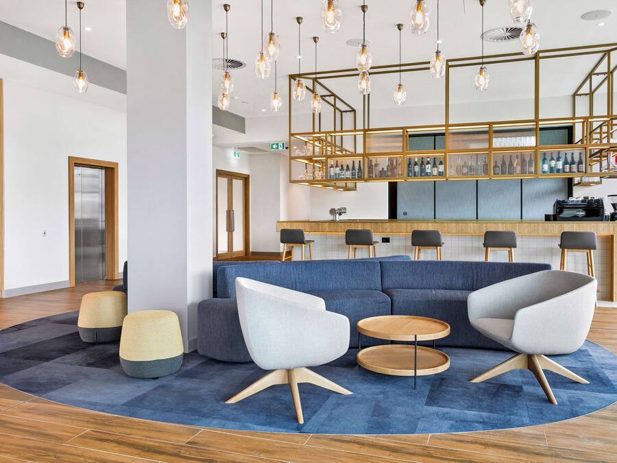 A modern lobby with a blue carpet, two white armchairs, a wooden coffee table, and a bar area with a glass display.
