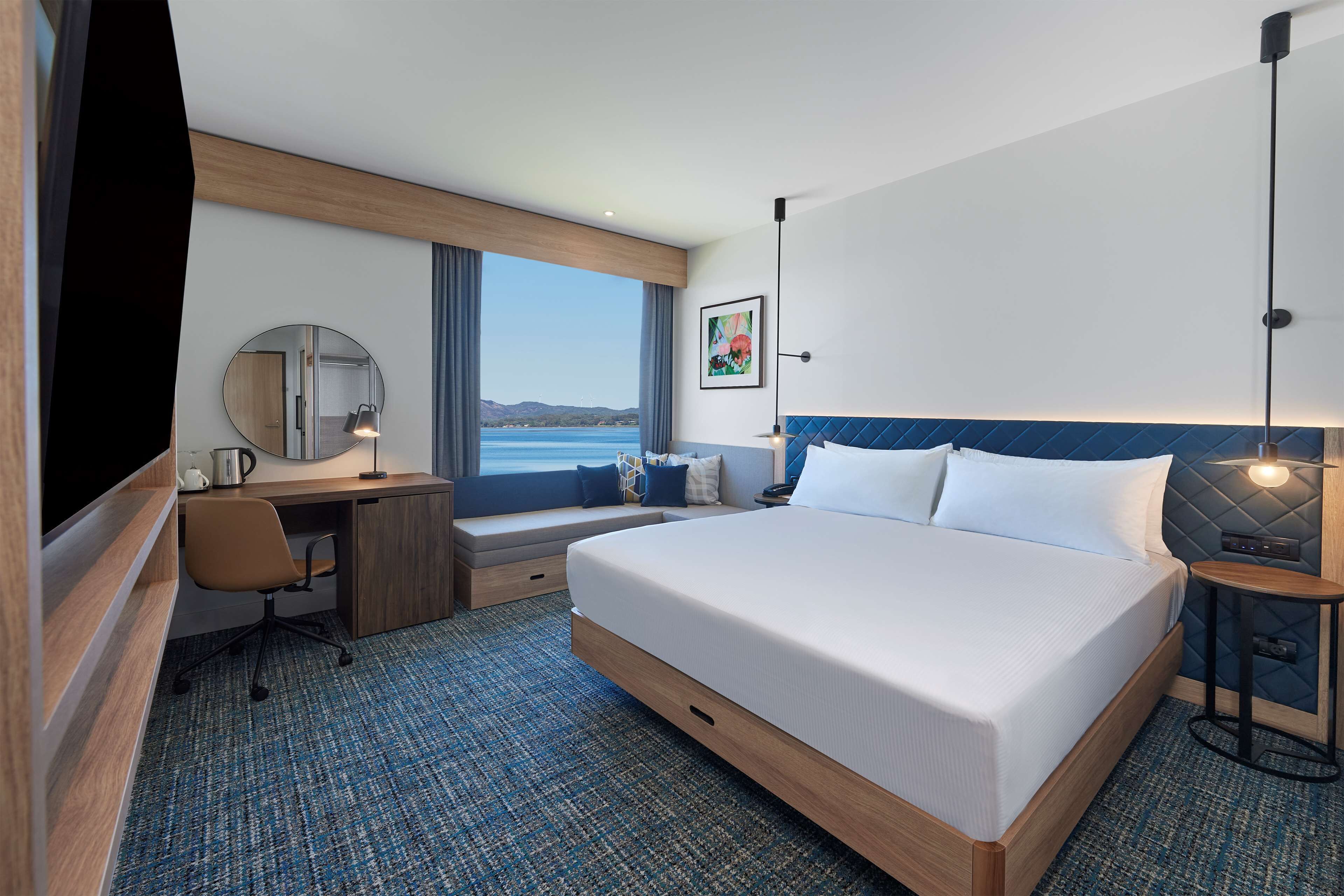 A hotel room with a large bed, a desk with a chair, a window with a view of the sea, and a painting on the wall.