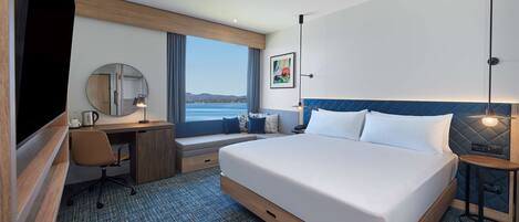 A hotel room with a large bed, a desk with a chair, a window with a view of the sea, and a painting on the wall.