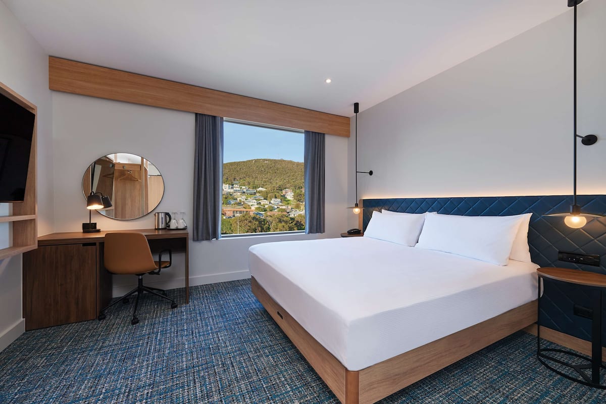 Ein Hotelzimmer mit einem großen Bett, einem Schreibtisch, einem Sessel, einem runden Spiegel und Blick auf eine Stadtlandschaft durch das Fenster.