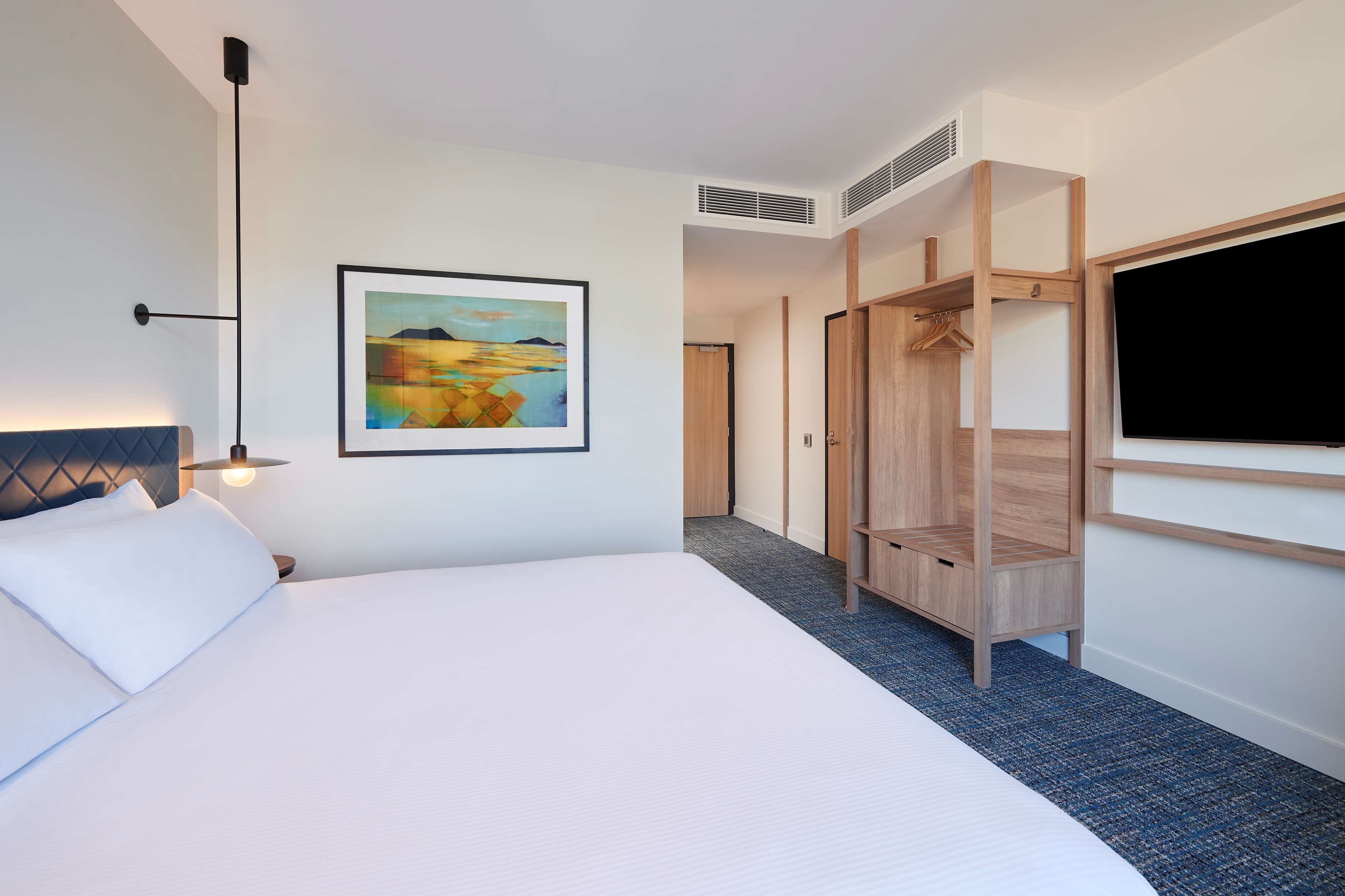 A hotel room with a large bed, a painting on the wall, a wardrobe, and a television.