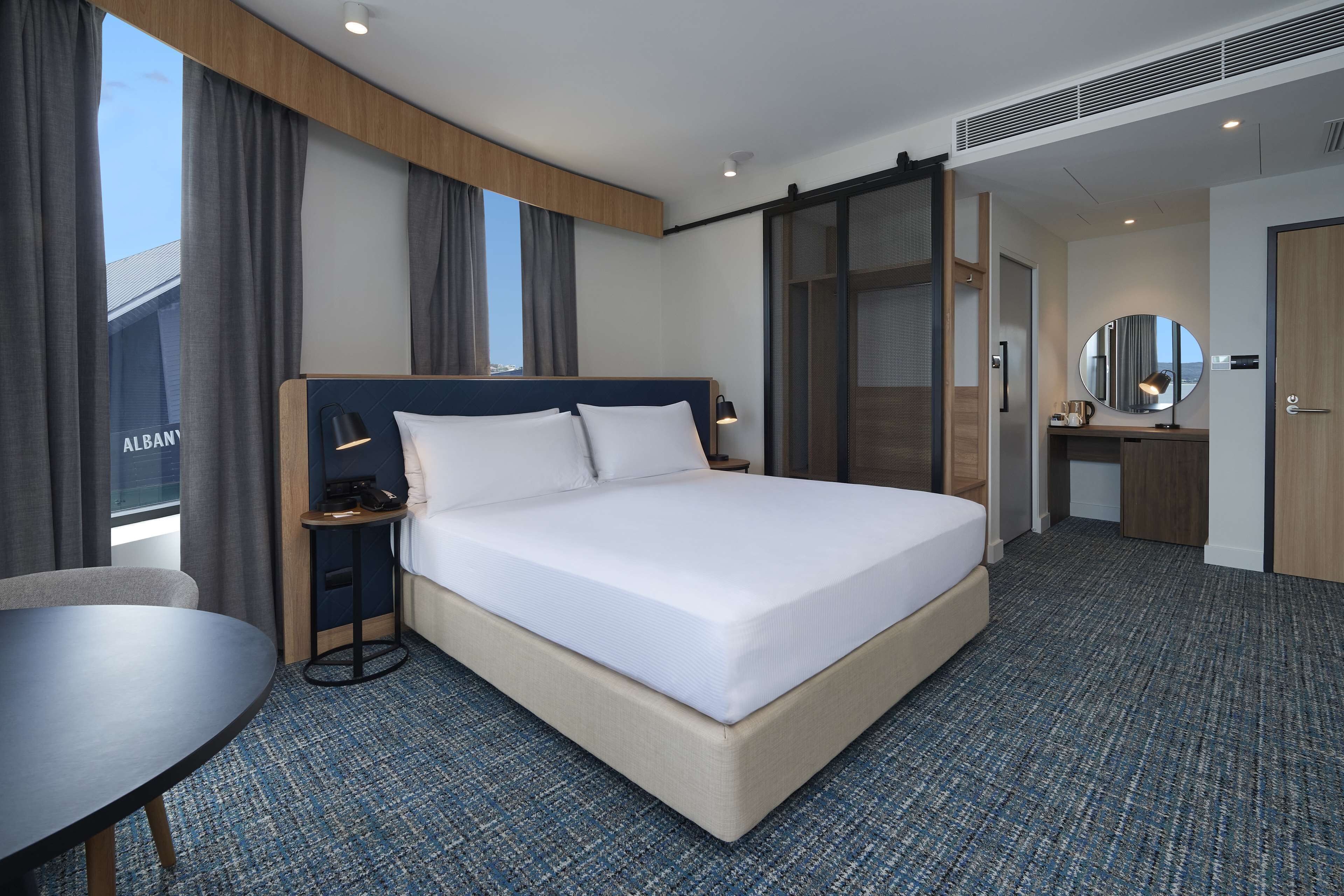 Habitación de hotel con una cama grande, un escritorio, una silla, una mesita, un espejo y vistas al exterior.