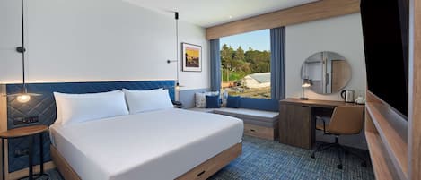 Habitación de hotel con una cama grande, un escritorio, una silla, una lámpara de escritorio, una mesita de noche, un espejo y una ventana con vistas a árboles.