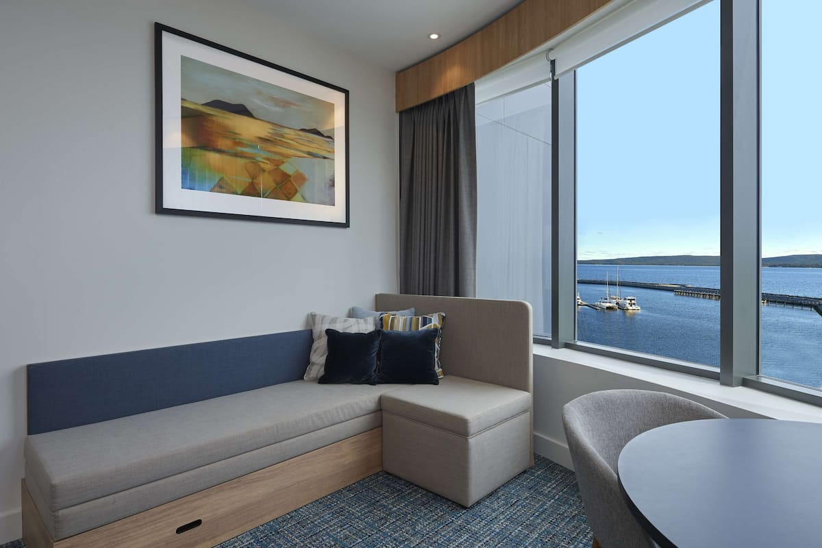 Ein Hotelzimmer mit einer Couch, einem Sessel, einem Tisch, einem großen Fenster mit Blick auf das Wasser und einen Steg sowie einem gerahmten Gemälde an der Wand.