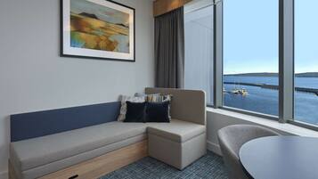A hotel room with a sofa, a chair, a table, a large window with a view of the water and a pier, and a framed painting on the wall.