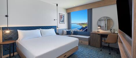 A hotel room with a large bed, a desk, a chair, a small stool, a desk lamp, a mirror, and a view of the sea.