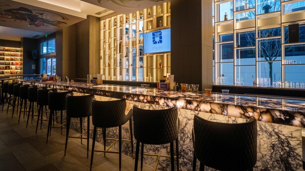 A bar area with a marble countertop, black cushioned bar stools, a television screen, and shelves with bottles. - Embassy Suites by Hilton Atlanta Midtown (Atlanta)