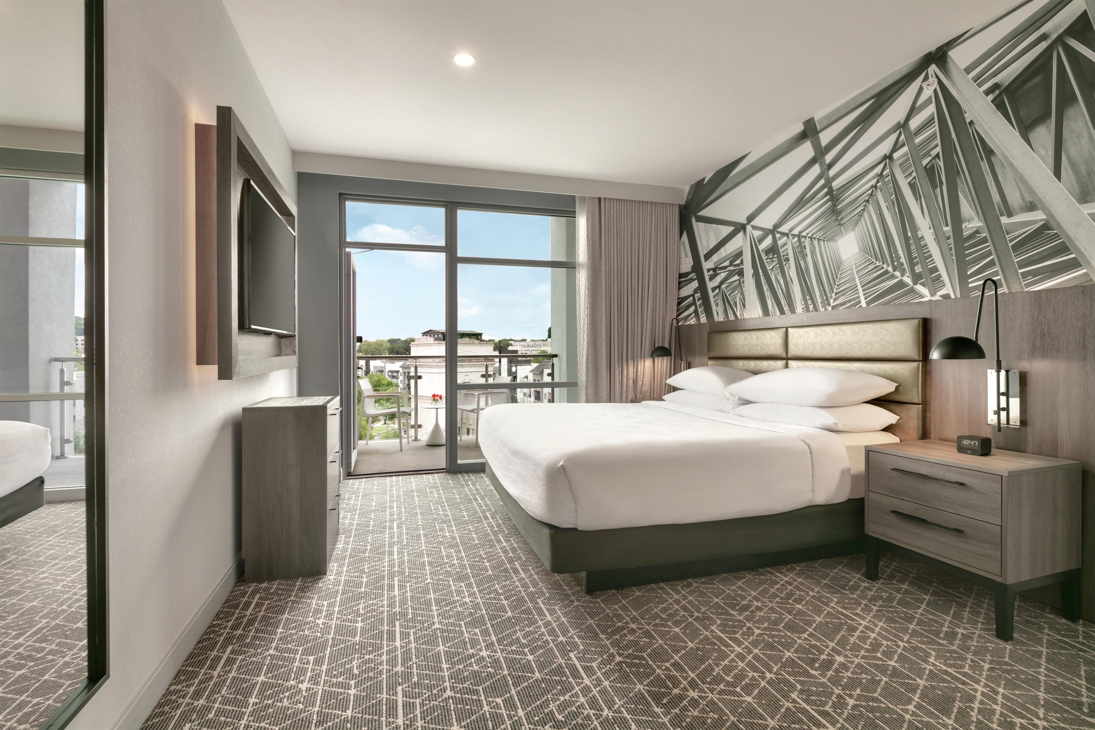 A hotel room with a large bed, a bedside table with a lamp, a view of the outdoors through a glass door, and a modern ceiling design.