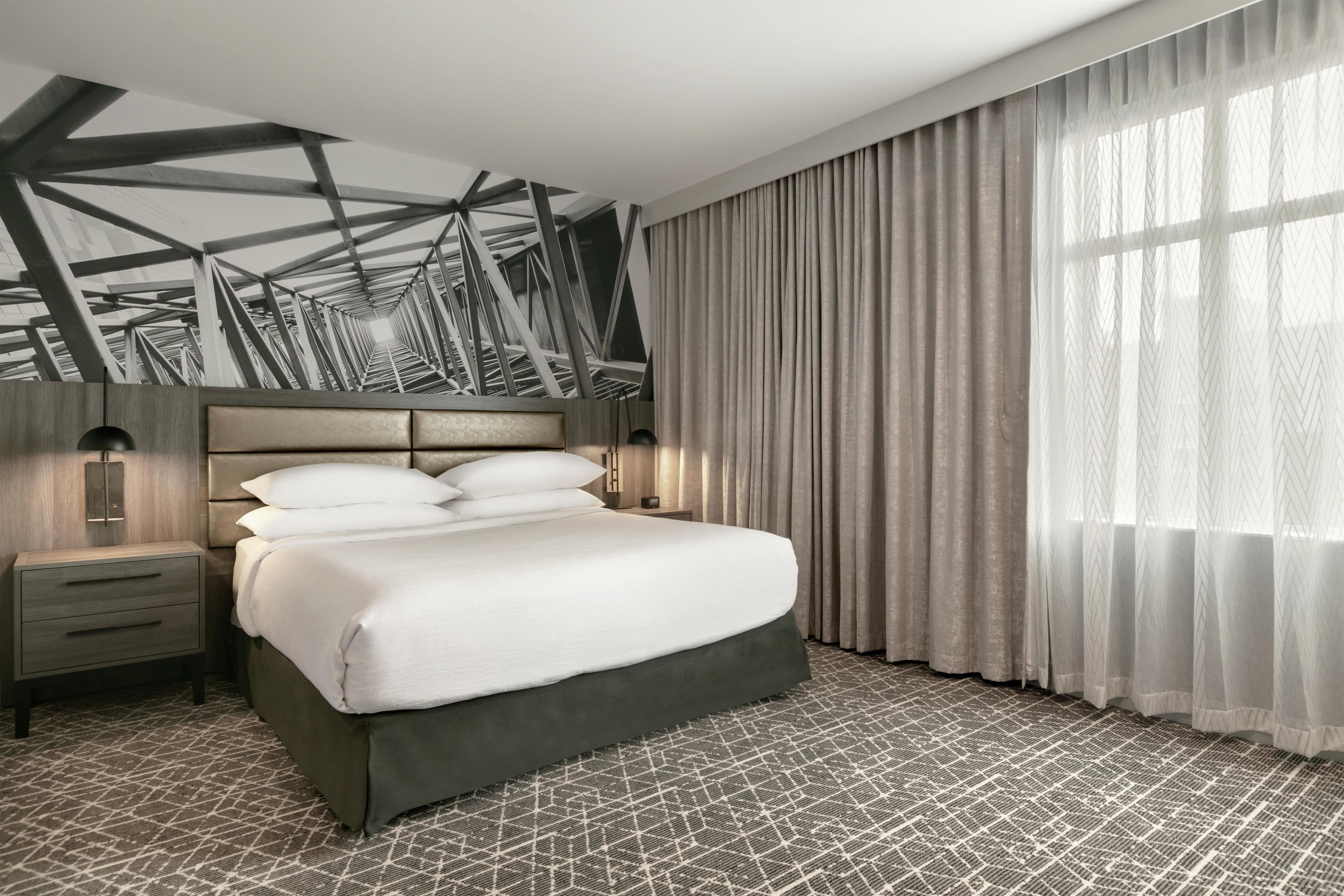 A modern hotel room with a large bed, a nightstand, a wall-mounted lamp, and a window with curtains.