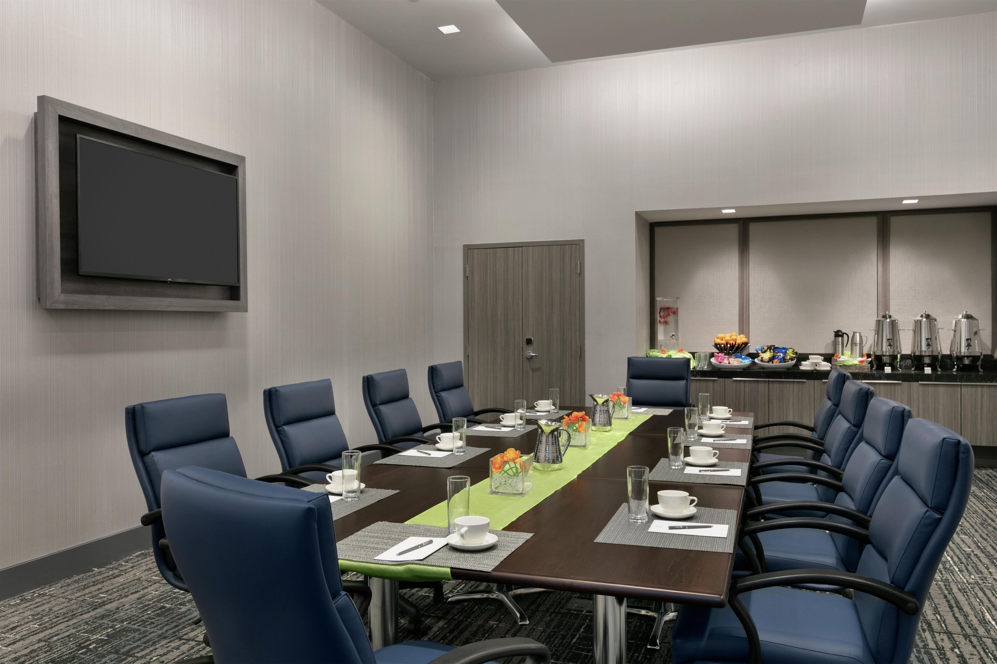 Un salón de conferencias con una mesa larga, sillas azules, un televisor de pantalla plana y una zona de kitchenette.
