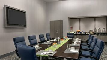 Un salón de conferencias con una mesa larga, sillas azules, un televisor de pantalla plana y una zona de kitchenette.