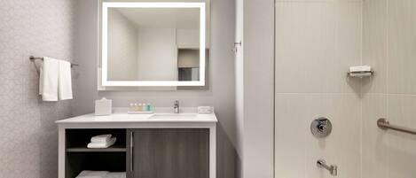 Une salle de bain moderne avec un meuble-lavabo blanc, un grand miroir, une douche équipée d’un pommeau de douche à main et un porte-serviettes.