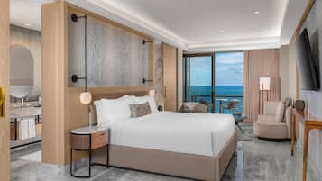 A modern hotel room with a large bed, a desk with a chair, a TV, and a view of the ocean.