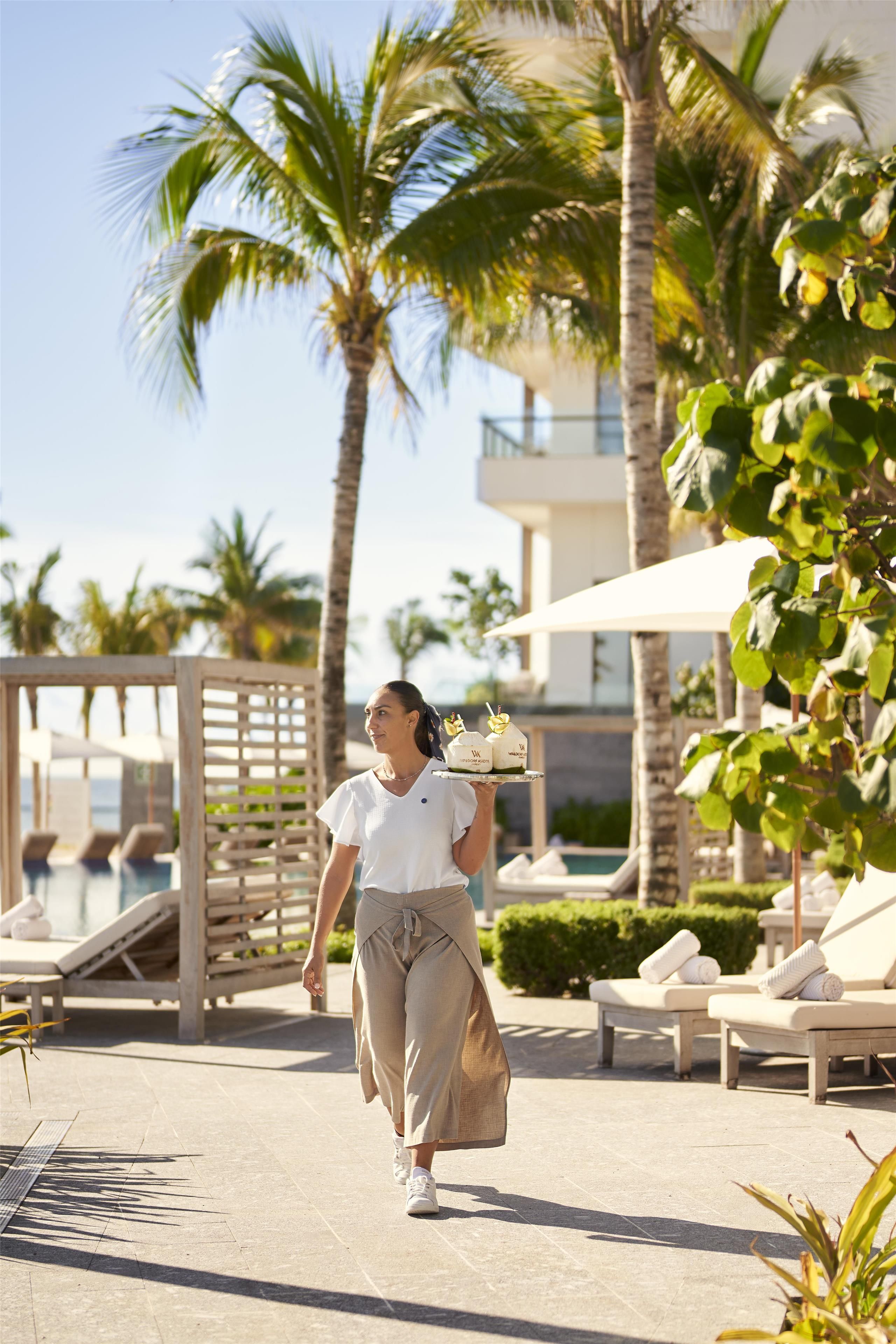 A person walking on a pathway with palm trees and lounge chairs, carrying a tray with drinks.