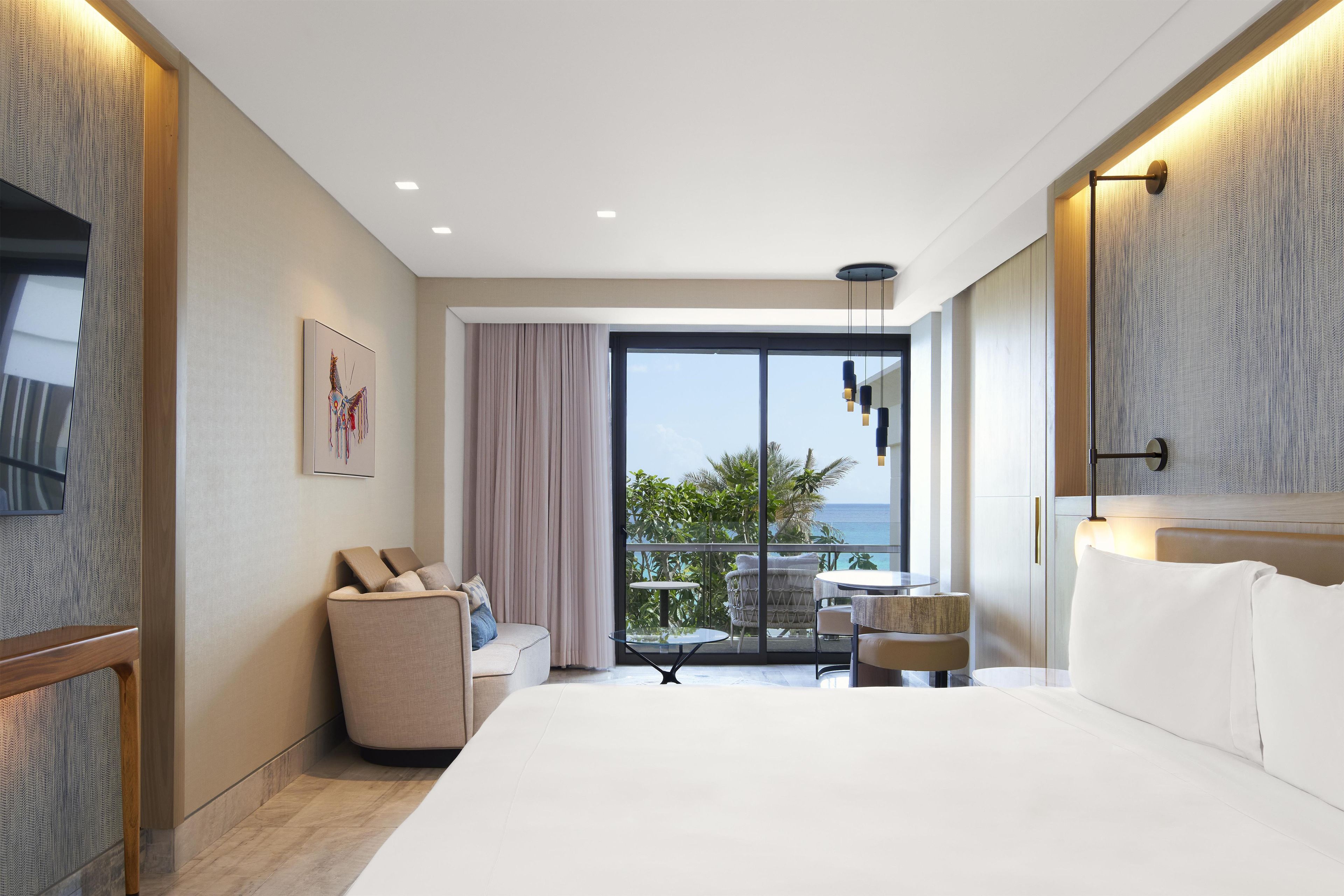 A modern hotel room with a large bed, a seating area with a table and chairs, a balcony with a view of the sea, and a large window.
