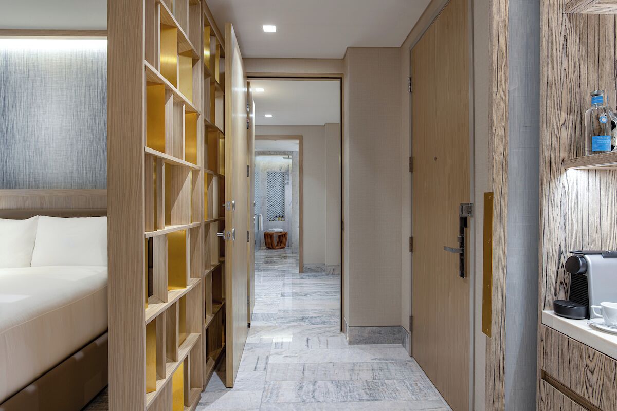 Un couloir d’hôtel avec un lit, des étagères en bois, une machine à café et, au fond, une salle de bain.
