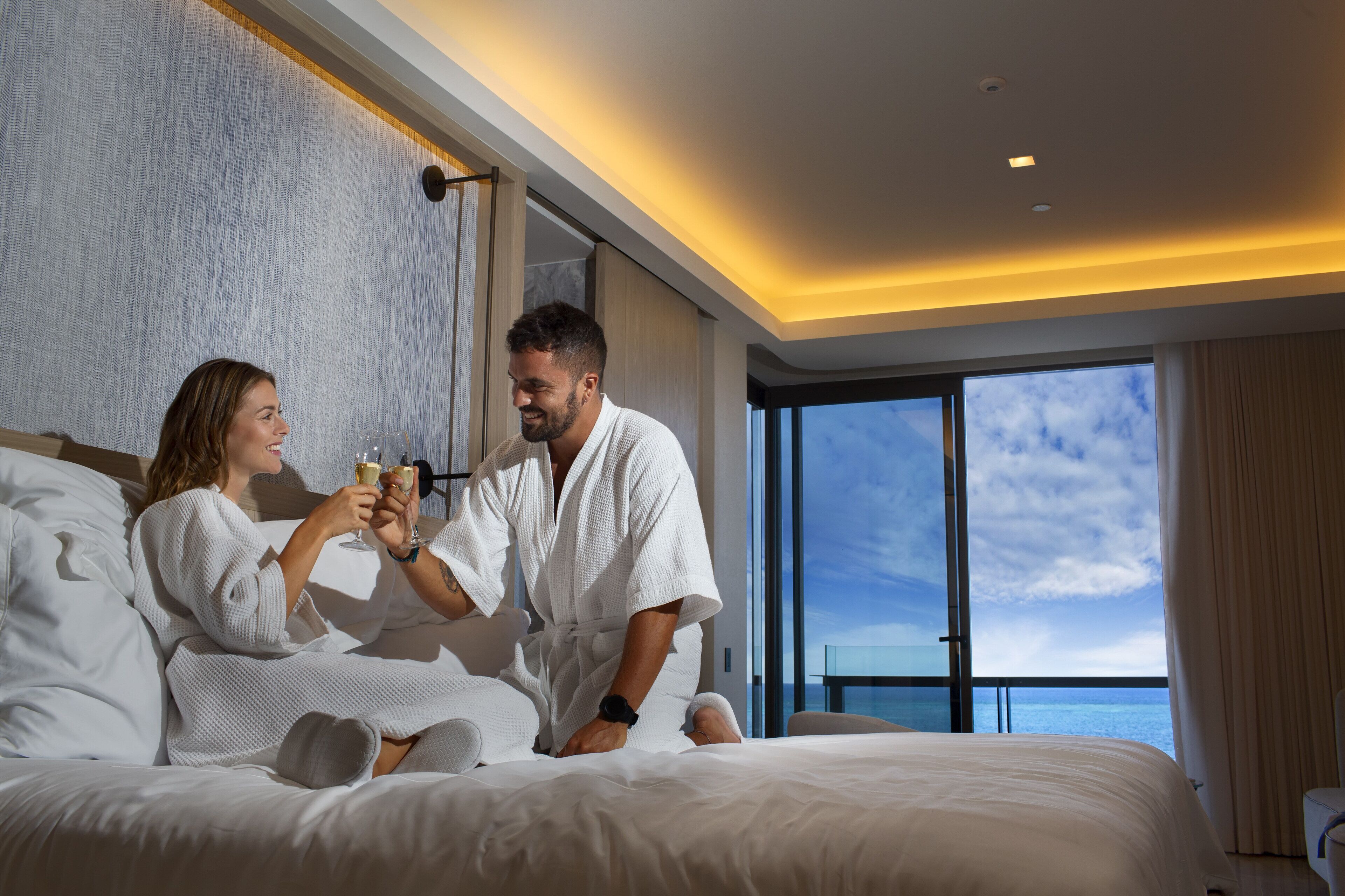 Una pareja en una habitación de hotel con una cama grande, vista al mar y un diseño interior moderno.