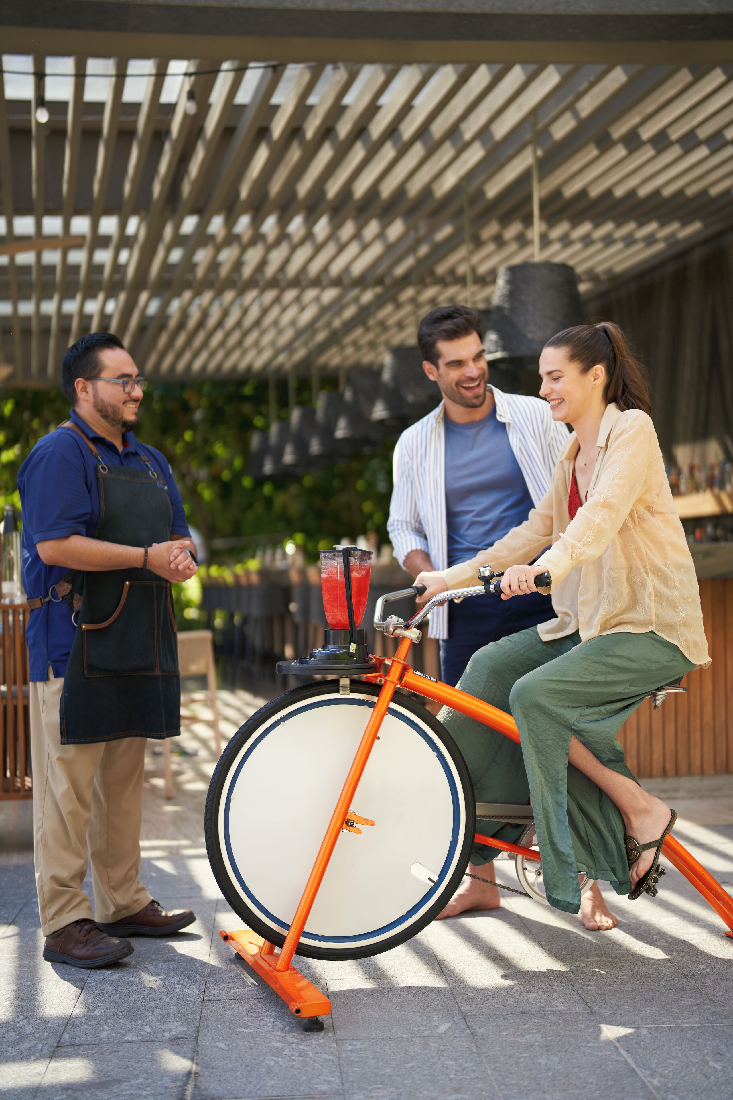 A person in a yellow shirt riding a stationary bicycle with a large wheel, another person in a blue shirt standing nearby, and a third person in a blue apron.