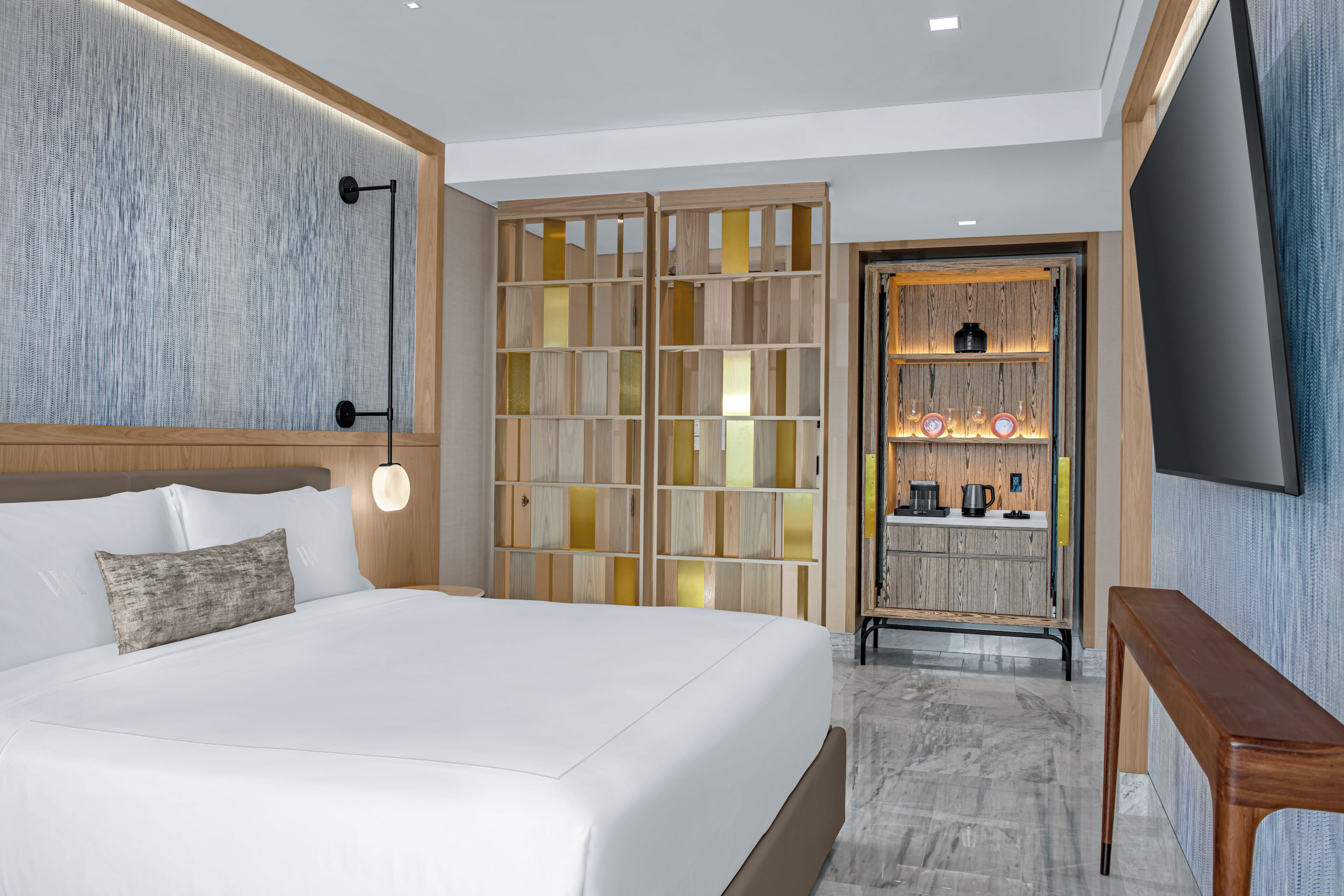 A modern hotel room with a large bed, a wooden wardrobe, a television, and a small kitchenette.
