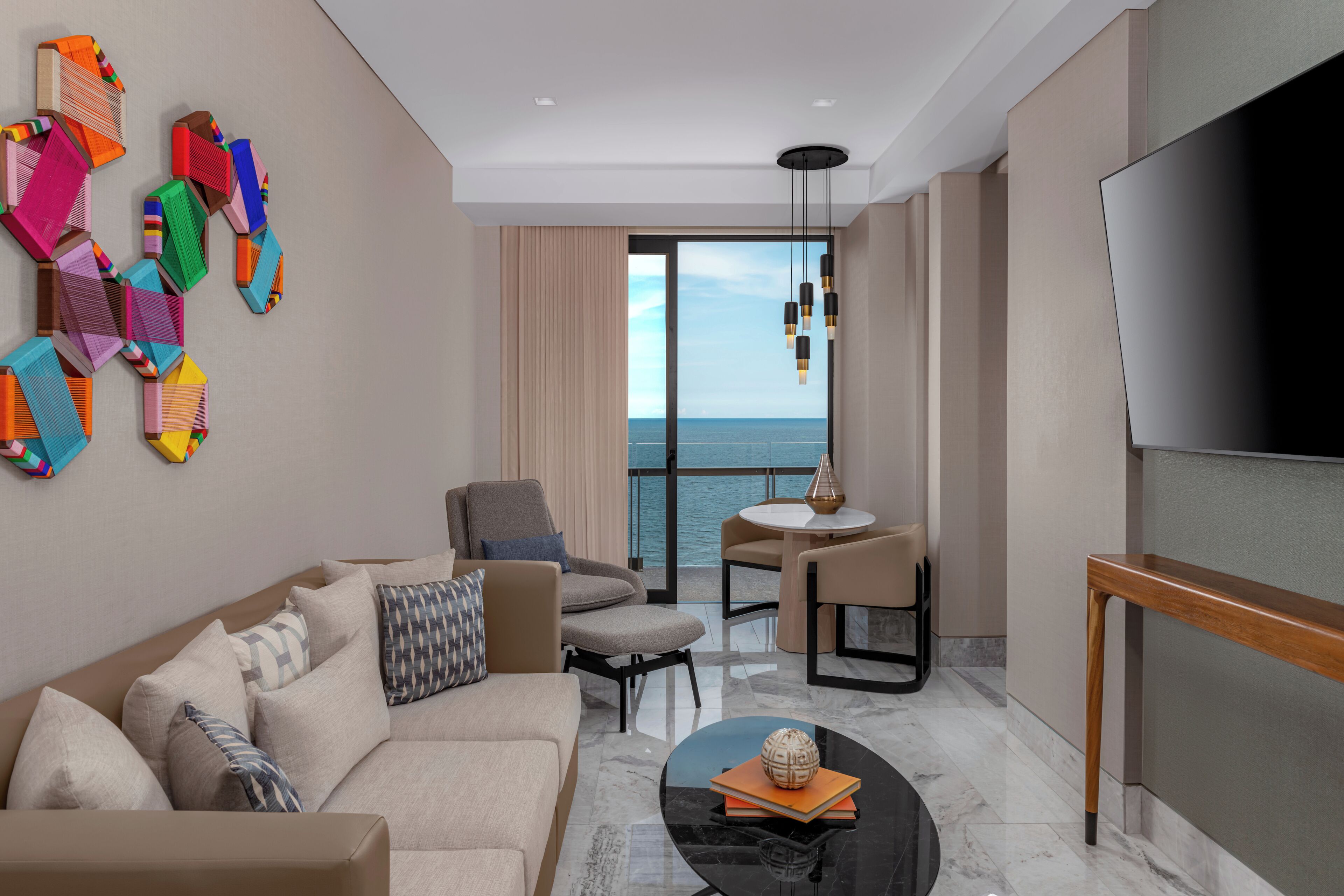 A modern hotel room with a sofa, armchairs, a coffee table, a TV, a balcony with a view of the sea, and a unique wall art installation.
