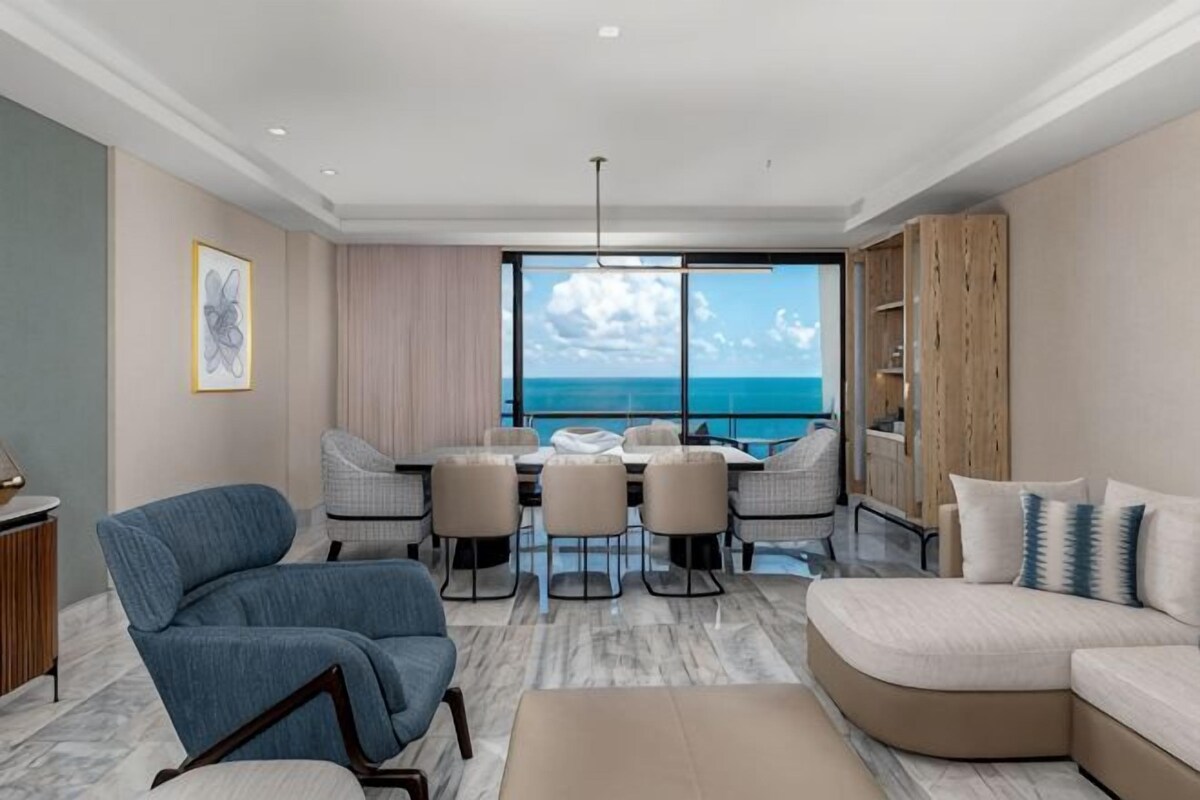 A modern living room with a dining area, a large window with a view of the sea, and a curved sofa.