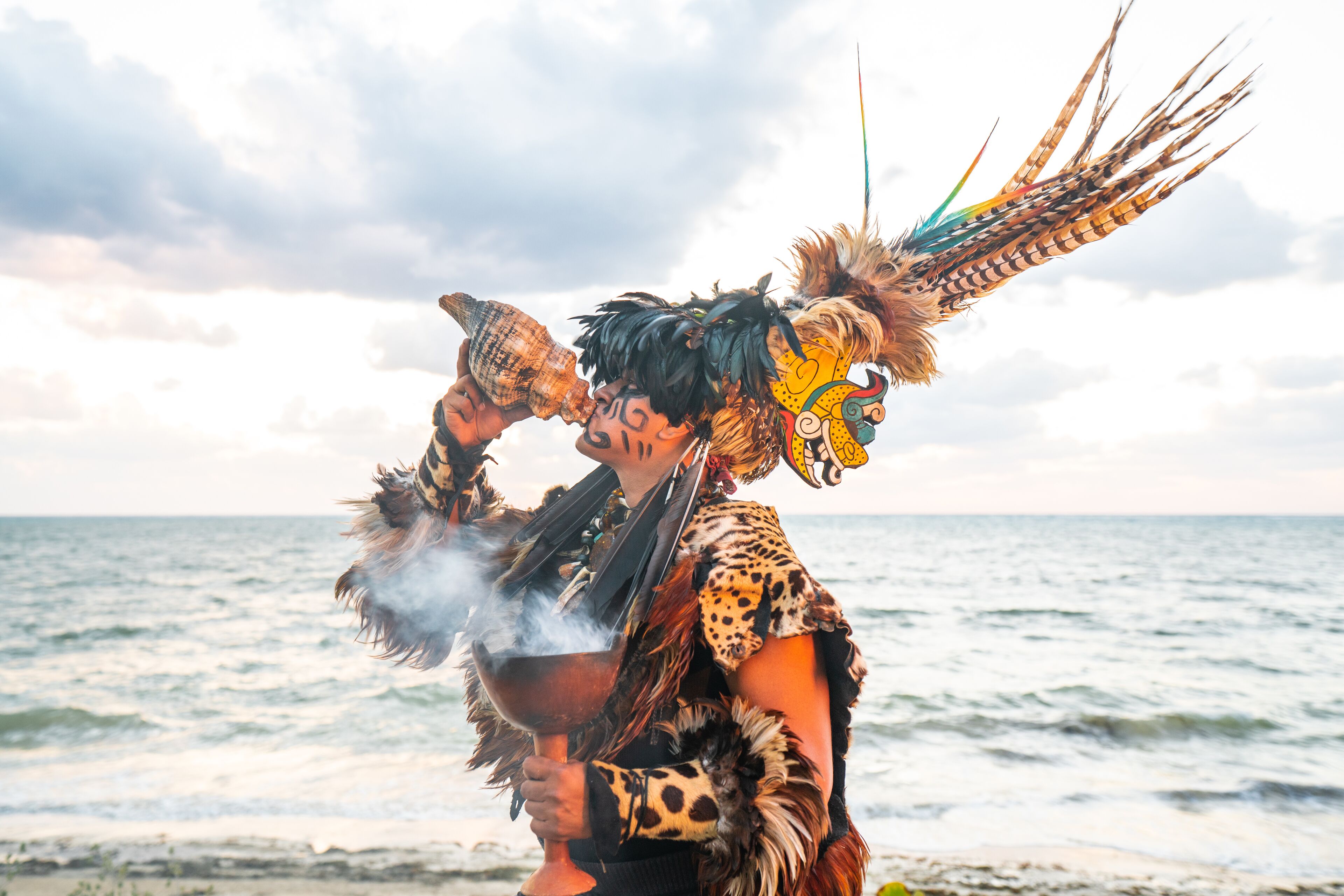 A person in a costume with feathers and a mask, holding a bowl and a cup, by the sea.