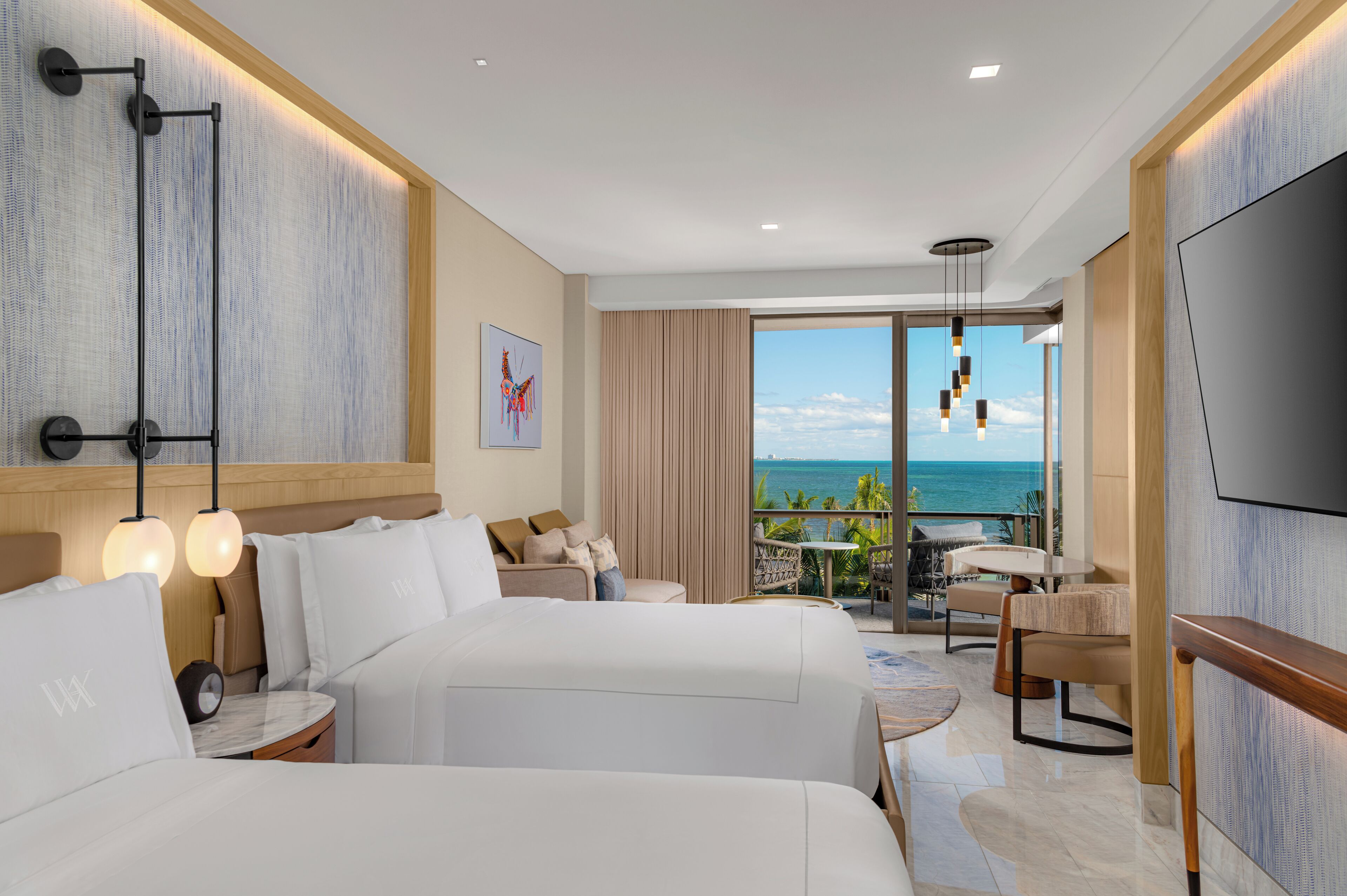 A hotel room with two beds, a balcony with a view of the ocean, a dining area, and a TV.
