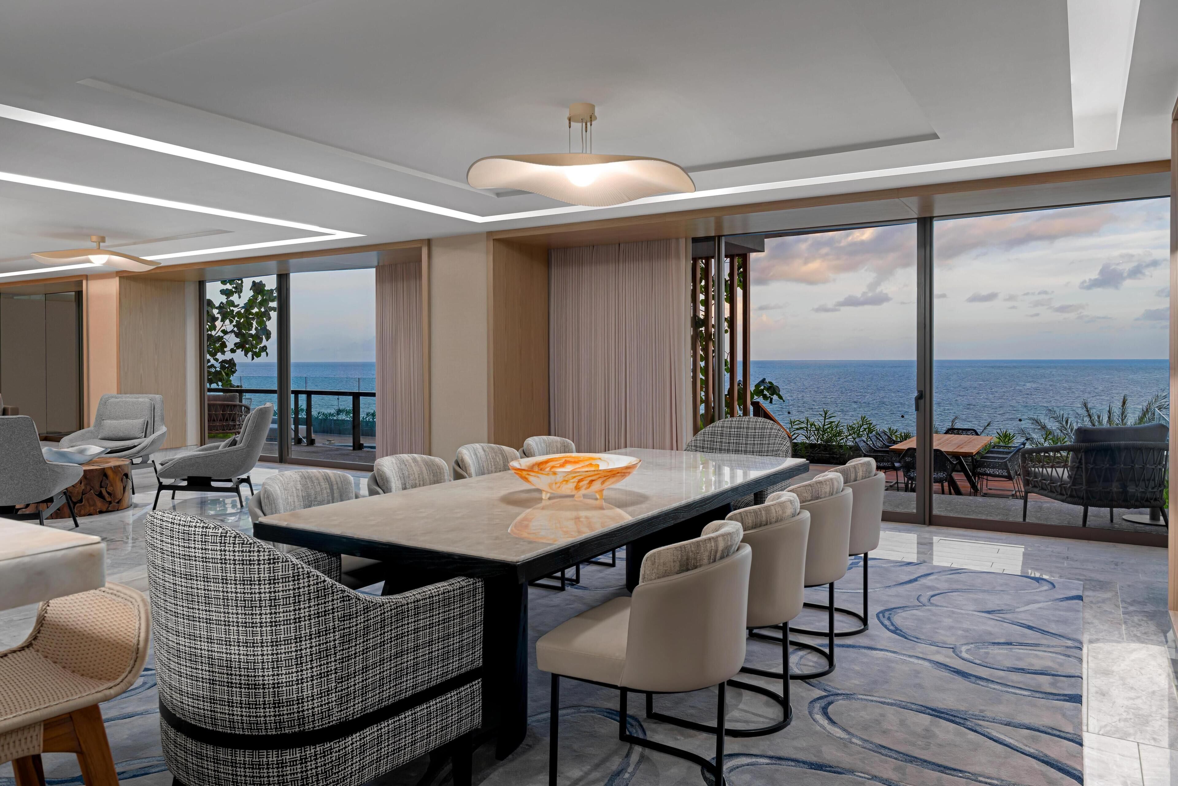 Ein moderner Essbereich mit großem Tisch und Stühlen, großen Fenstern mit Meerblick und einer Decke mit Einbaustrahlern.