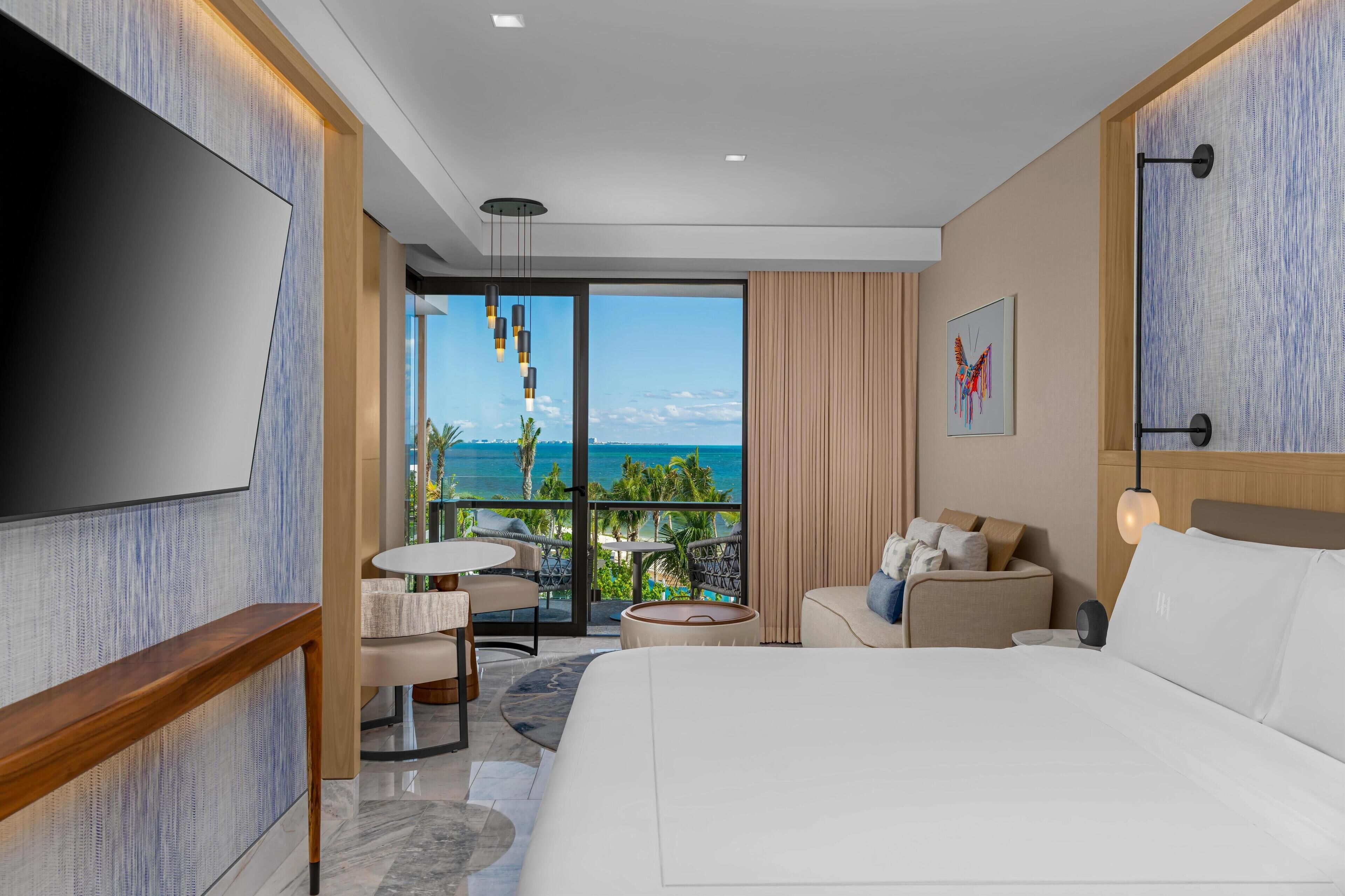 A modern hotel room with a large bed, a TV, a balcony with a view of the ocean, and a small seating area with a table and chairs.