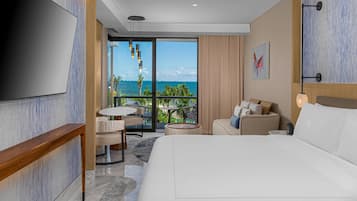 A modern hotel room with a large bed, a TV, a balcony with a view of the ocean, and a small seating area with a table and chairs.