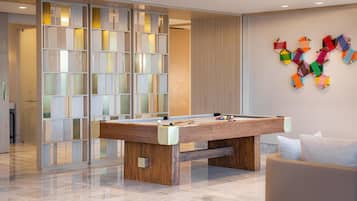 Uma mesa de bilhar moderna em ambiente interno, com acabamento em madeira, divisória de vidro com painéis jateados e uma obra de arte abstrata geométrica na parede.