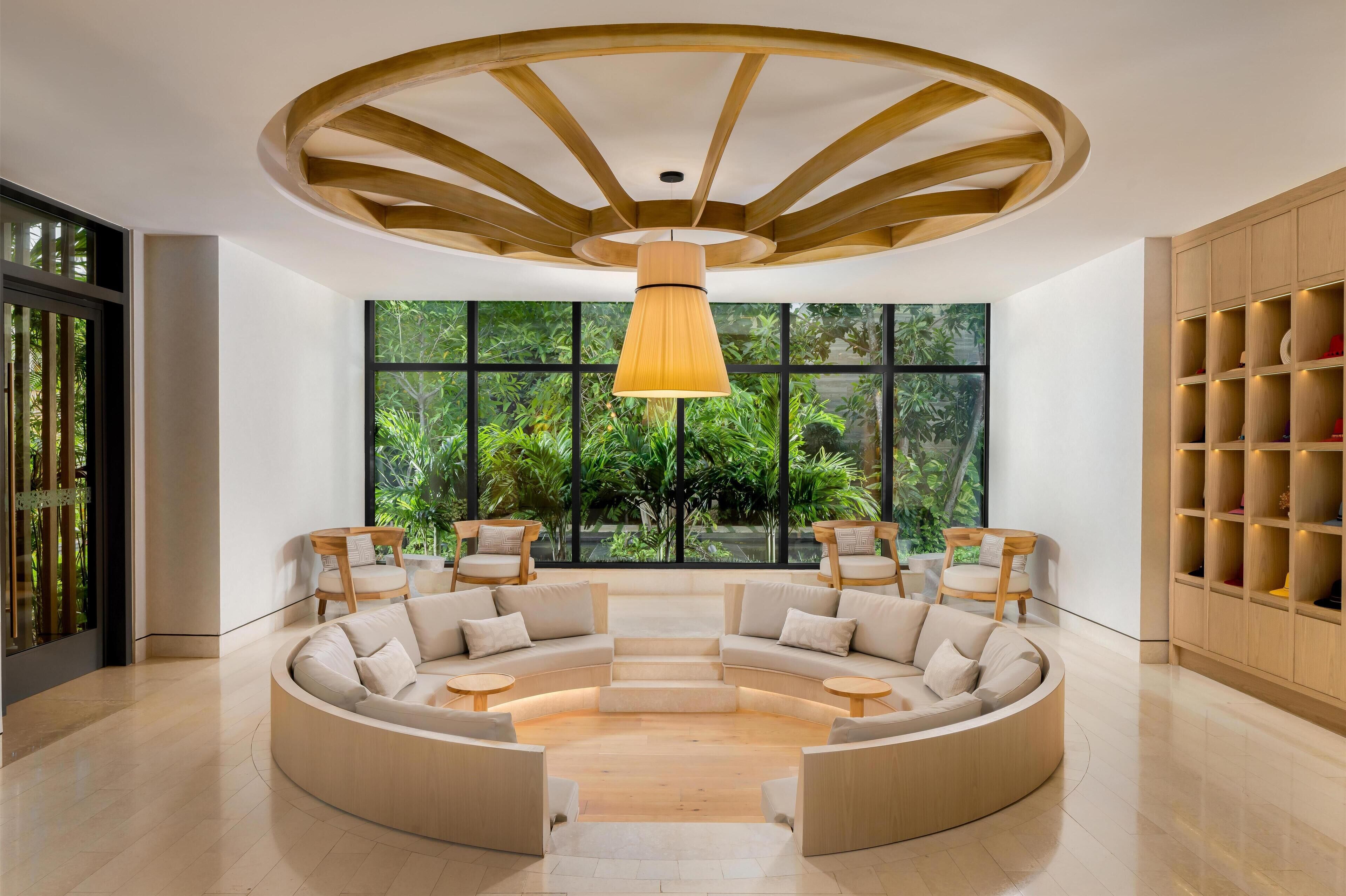 A modern interior with a circular seating area, large windows with a view of greenery, and a wooden ceiling structure.