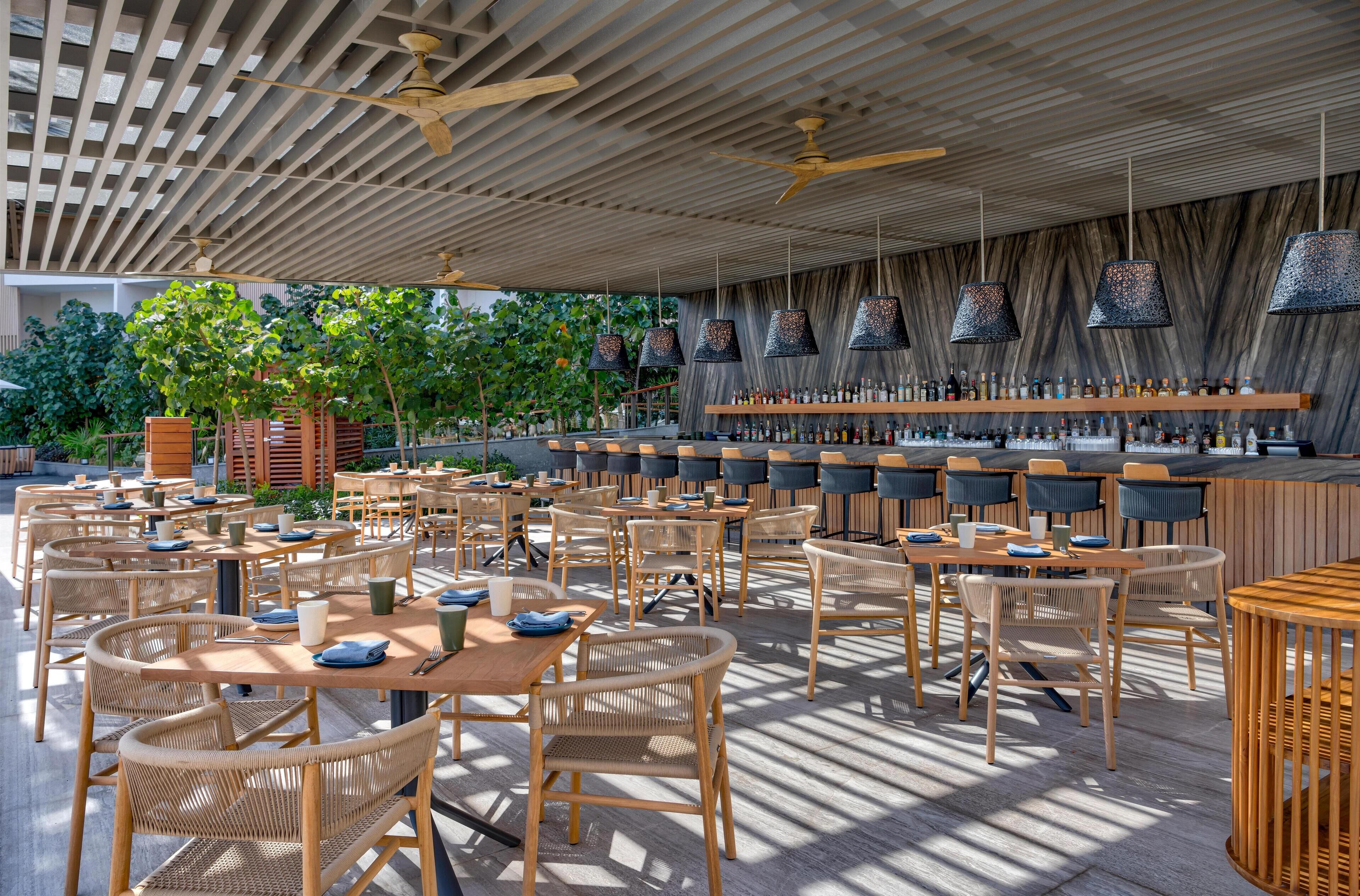 Un espace de restauration en plein air avec des tables et des chaises en bois, un bar doté d’étagères remplies de bouteilles et un plafond orné de lattes de bois et de lampes suspendues.