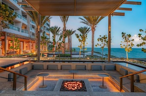 A modern outdoor seating area with a fire feature, palm trees, and a view of the ocean.