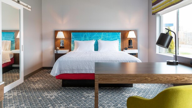 Una habitación de hotel con cama, mesitas de noche, escritorio, silla, lámpara y ventana.