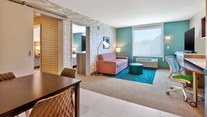 A modern hotel room with a dining area, a sofa, a desk, and a large window. - Home2 Suites by Hilton Grand Blanc Flint (Grand Blanc)