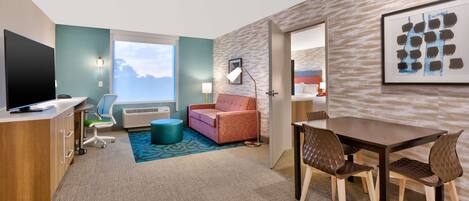 A hotel room with a desk, a television, a sofa, a dining table with chairs, a window with a view, and a wall-mounted artwork.