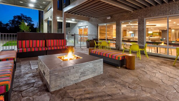 An outdoor patio area with a fire pit, seating, and a view of the building's interior. - Home2 Suites by Hilton Grand Blanc Flint (Grand Blanc)