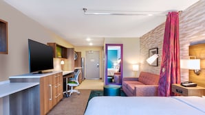 A hotel room with a bed, desk, TV, and a bathroom visible through an open door. - Home2 Suites by Hilton Grand Blanc Flint (Grand Blanc)