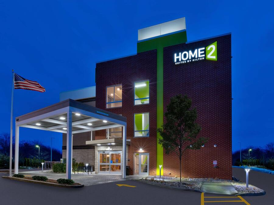 Entrada de un hotel con un área de espera techada, un letrero llamativo de "HOME 2 Suites by Hilton" y una bandera estadounidense.