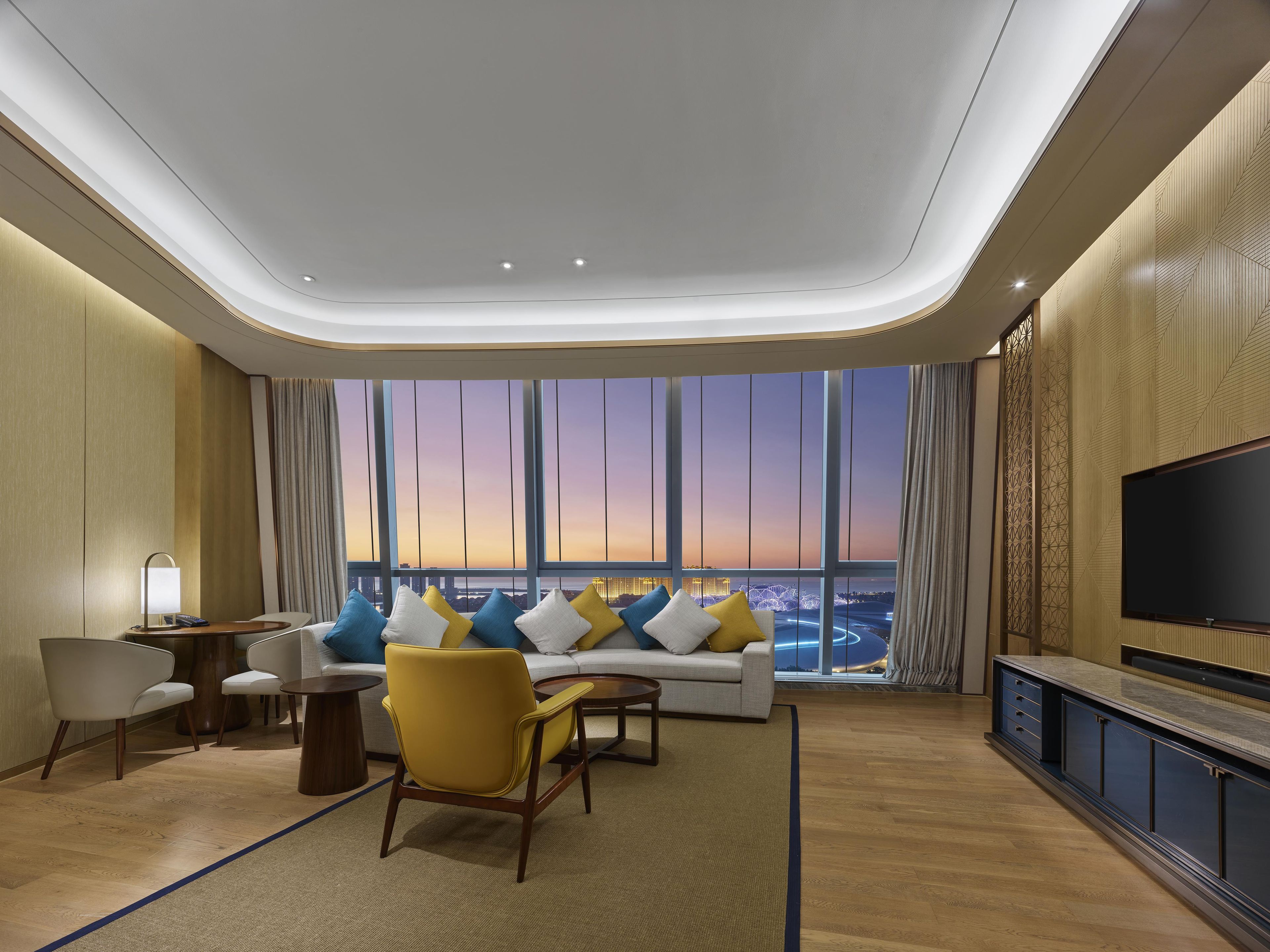 A modern hotel room with a large window, a sofa with cushions, a television, and a wooden floor.