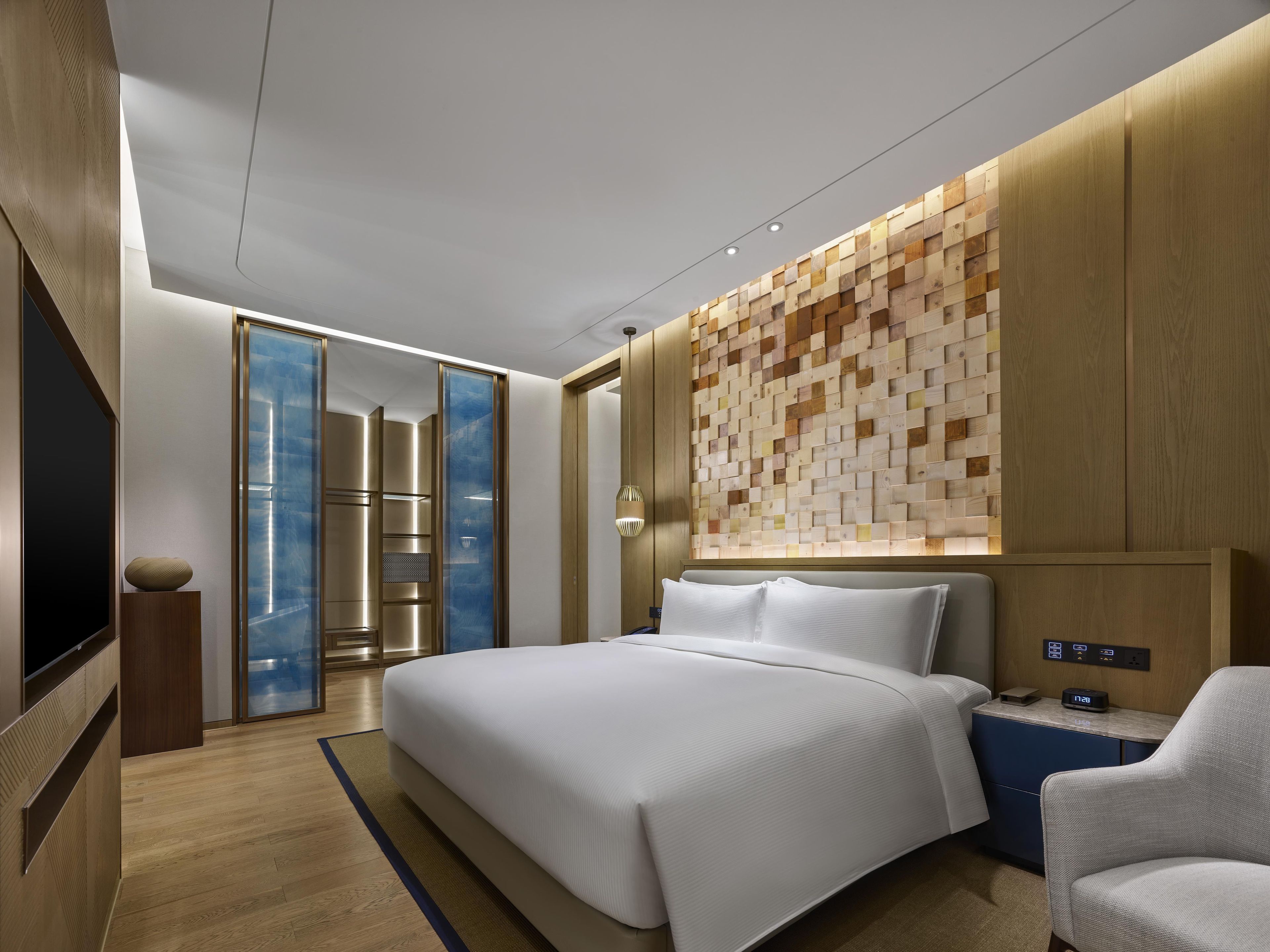 A modern hotel room with a large bed, a TV, a desk with a clock, and a wall with a geometric pattern.