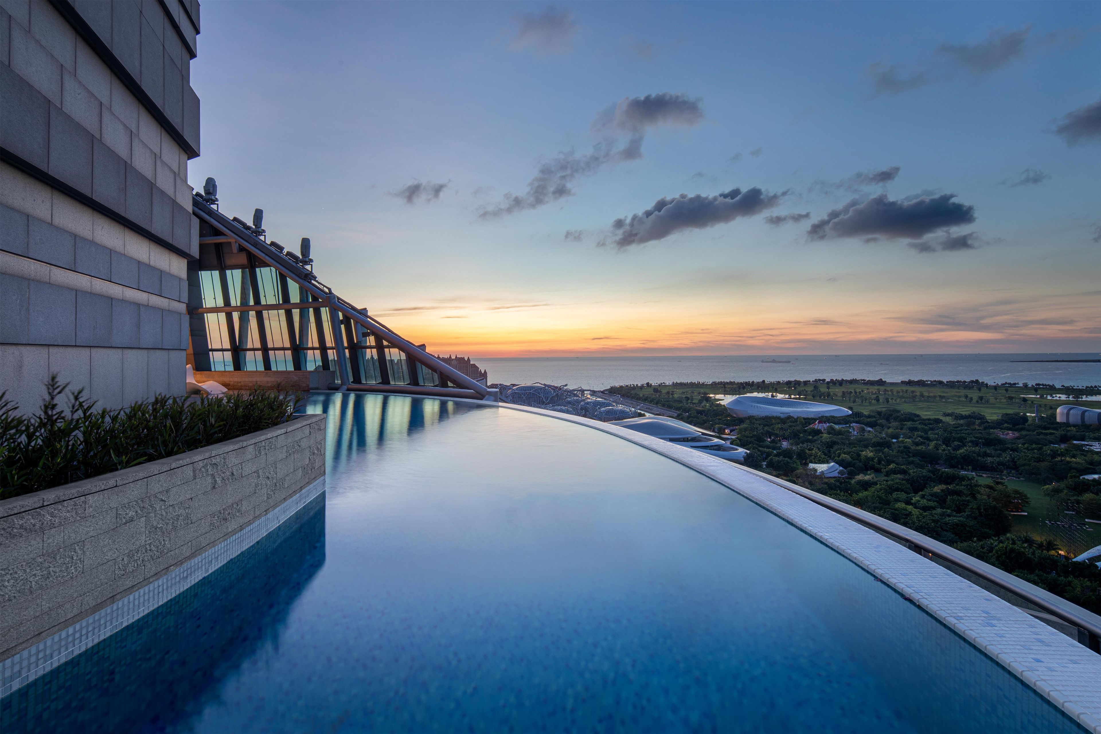 A modern hotel with a long swimming pool, a glass bridge, and a view of the city at sunset.